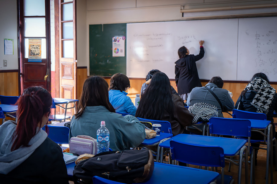 En la imagen aparece una sala de clases cojn estudiantes que ponen atención a la profesora que escribe en el pizarrón