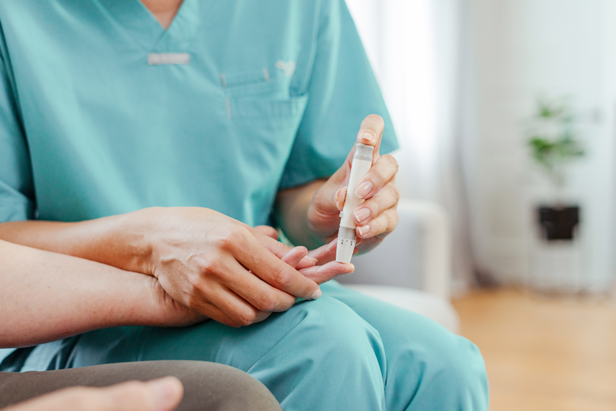 Blurred image of a healthcare professional in a turquoise gown holding a finger and a medical device, possibly a lancet, to measure blood sugar levels.
