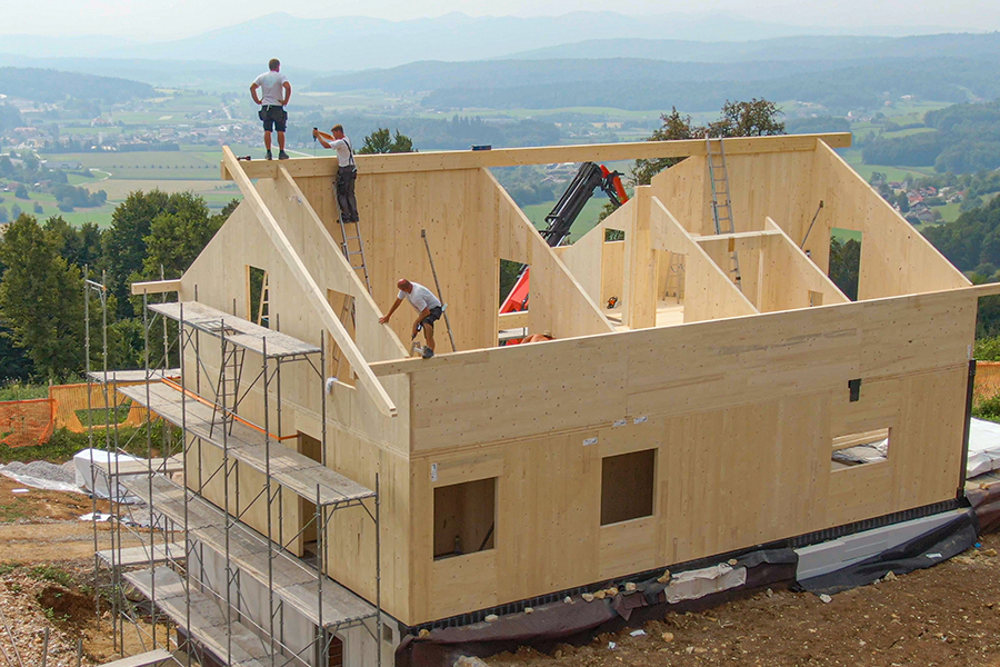 Photograph of construction workers on the roof of a large wooden house under construction.