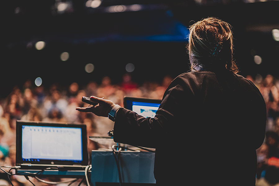 Photograph of a speaker behind a podium with two laptops, gesturing to a large audience.
