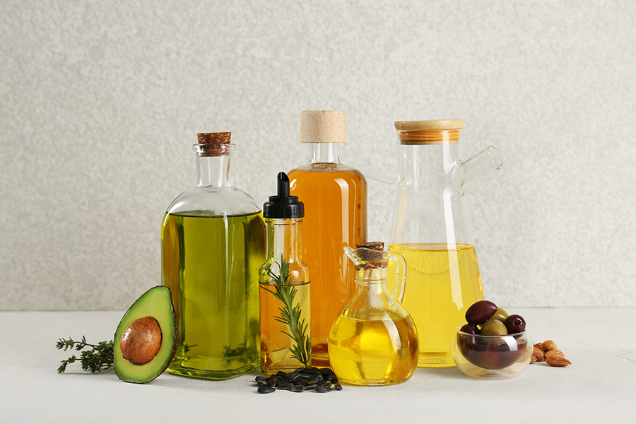Una variedad de aceites se exhiben en diferentes botellas de vidrio, junto con un aguacate cortado por la mitad, aceitunas y almendras, sobre una superficie de color claro.