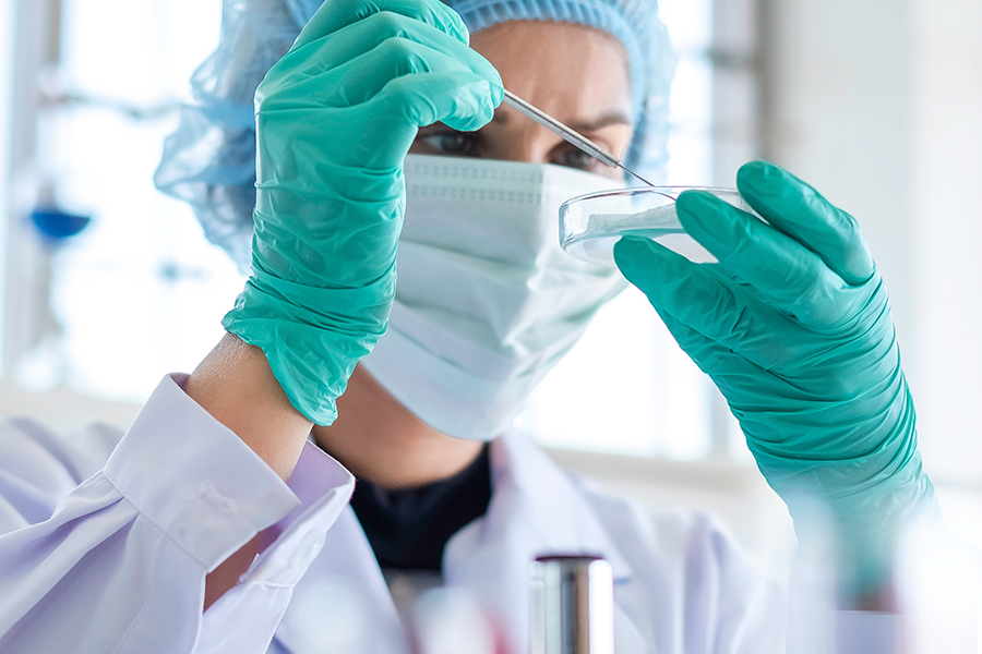 A blurrrd photo shows a woman in a lab coat and face mask looking intently at a dropper she is holding in her gloved hands.