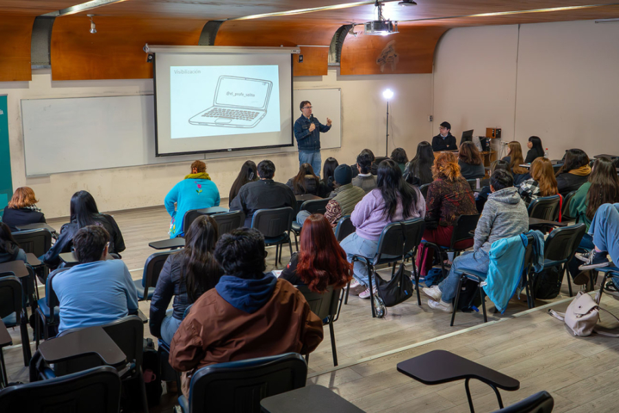 Docente del programa se dirige a la audiencia que está sentada en las sillas de la sala en donde se realizó el evento. De fondo, una pantalla gigante en donde aparecen las informaciones de la charla.