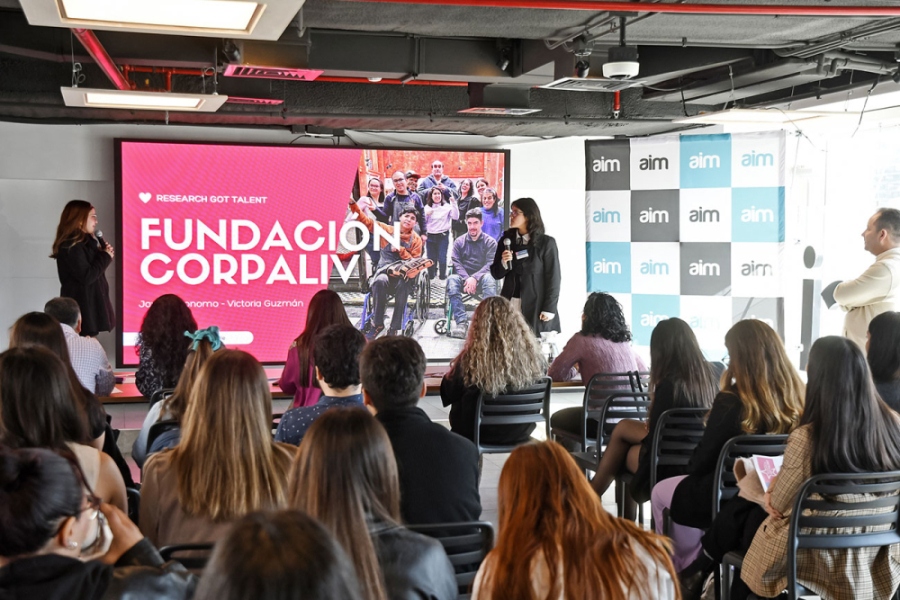 Fotografía de la actividad en la que aparece el público sentado mirando hacia el escenario. Dos personas le hablan a los asistentes y fondo una pantalla con el nombre del evento en color rojo con letras blancas.