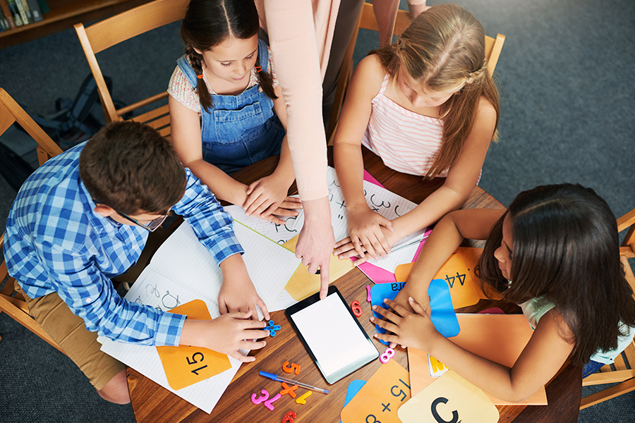An overhead shot captures four young students and an adult's hand pointing to a digital tablet while they work on a mathematics lesson surrounded by number cards, papers with equations, and colorful manipulatives on a wooden table.