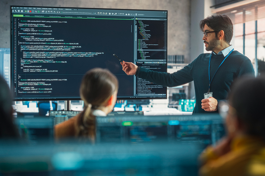 A professor points at a large screen displaying lines of computer code while teaching a class of students in a technology educational setting.