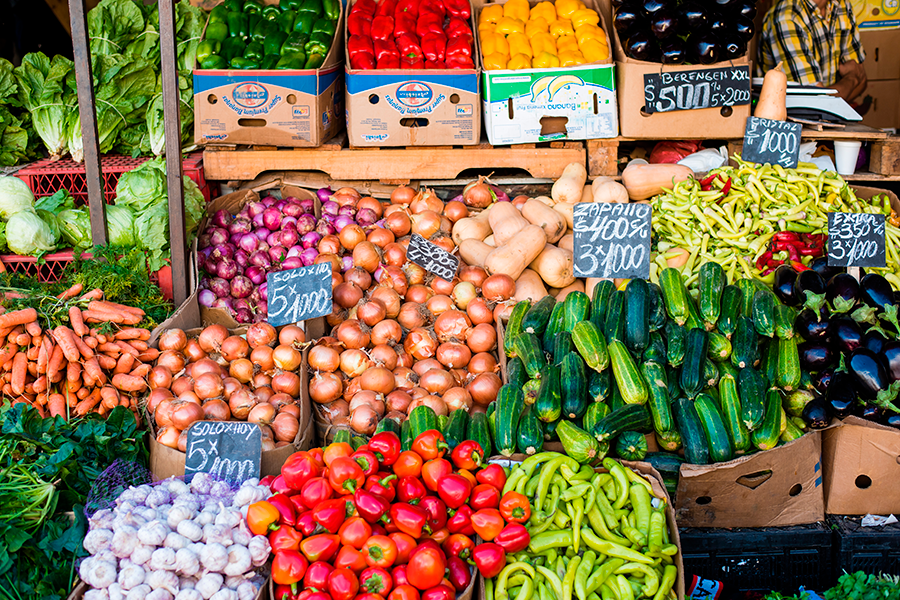 This photograph captures a vibrant, abundant display of fresh produce at a street market, featuring piles of colorful peppers, onions, carrots, squash, garlic, and cucumbers, many with handwritten price signs in Spanish.
