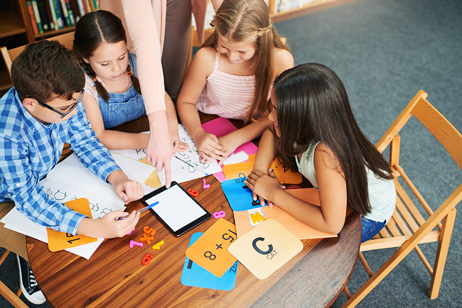 Um professor aponta para um tablet enquanto quatro alunos trabalham em atividades matemáticas e de aprendizagem em torno de uma mesa em uma sala de aula.