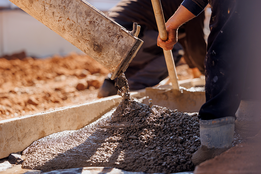 Un primer plano captura a trabajadores de la construcción vertiendo concreto húmedo y granulado de un balde sobre una base plana, con tierra seca visible en el fondo.