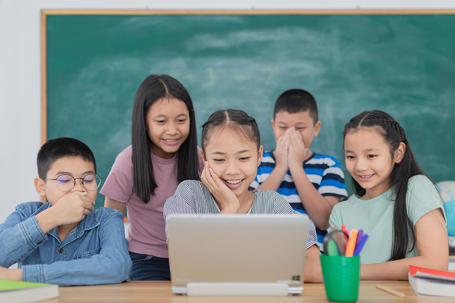 Five curious students gather around a laptop in a classroom, smiling while collaborating on a digital project together.