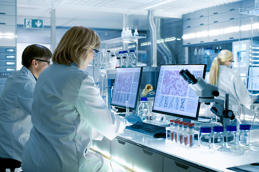 Científicos con batas blancas trabajan en la visualización de datos de células y ADN en un laboratorio luminoso y moderno, rodeados de microscopios y tubos de ensayo.