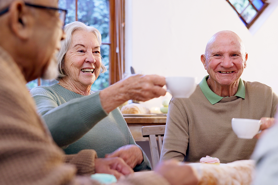 Three smiling seniors in warm light enjoy tea around a table, with a woman laughing while holding up her cup.