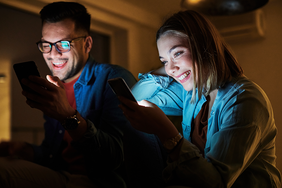 A smiling couple sits closely in a dark room, illuminated only by the light from the screens of the smartphones they are looking at.