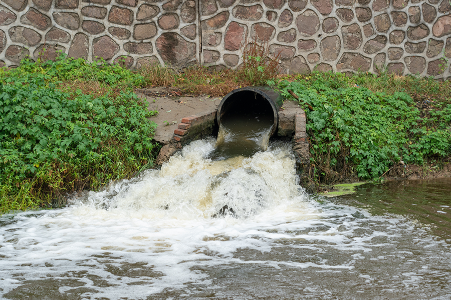 A black pipe protrudes from a stone wall and discharges a powerful jet of foamy, muddy water into a murky river surrounded by green algae.