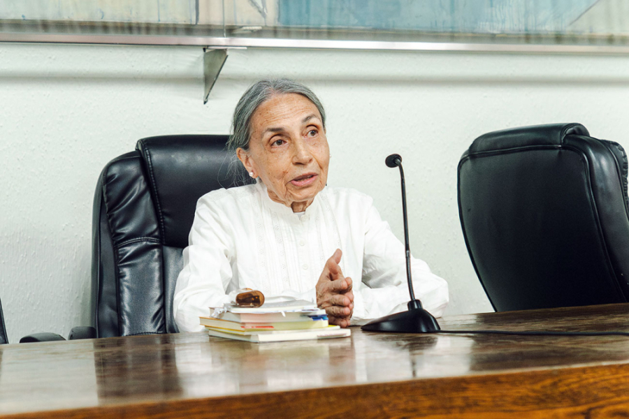 Imagen muestra a mujer exponiendo sentada sobre una silla negra y frente a un micrófono ubicado sobre una mesa