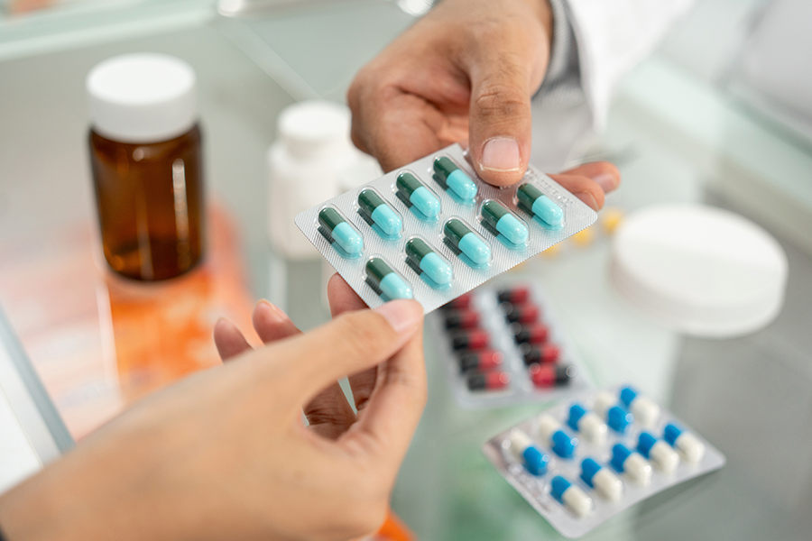 Close-up of two people's hands exchanging a silver blister pack of teal and dark green medicine capsules over a glass counter.