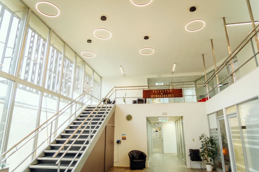 En la imagen aparece el hall de acceso a la Facultad de Humanidades. Una escalera a mano izquierda y oficinas a la derecha. Todo en fondo blanco y mucha luz de los grandes ventanales que dan a los patios de ciencia.
