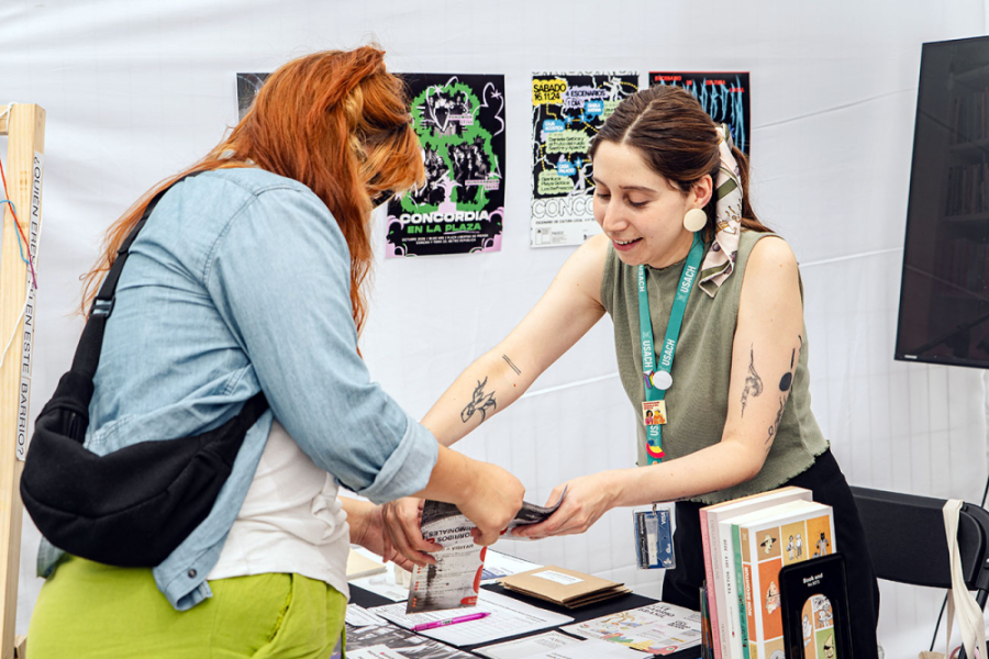Funcionaria VIME que atiende stand enseña material de trabajo a persona que visita la muestra