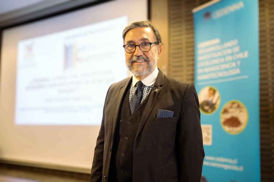 En la fotografía aparece el Dr. Zagal vestido con un terno negro y camisa a cuadrillé azul y blanco. De fondo, unas diapositivas de sus estudios. 