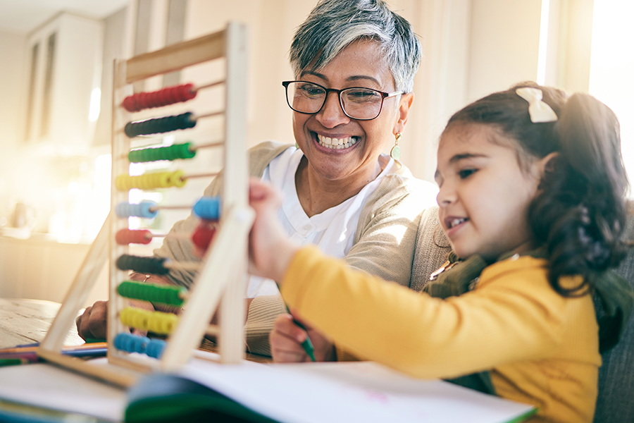 A smiling woman helps a young girl use a colorful wooden abacus. for learning at a table.