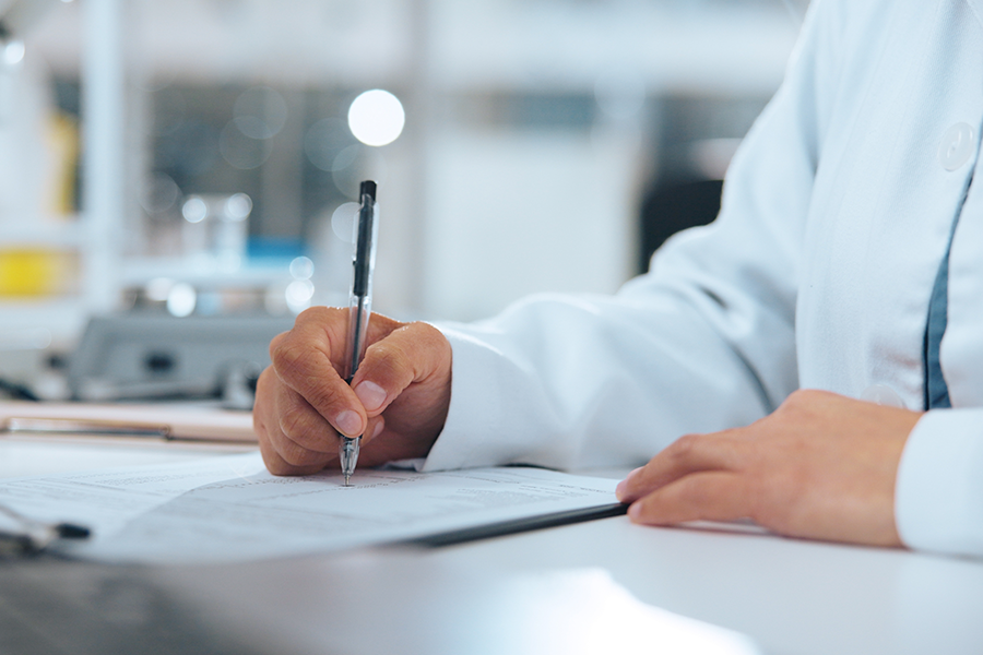 Close-up of a person in a white lab coat carefully writing notes on a document with a black pen.