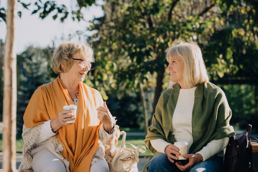 Dos mujeres sentadas en un banco del parque conversando mientras sostienen tazas de café a la luz del sol.