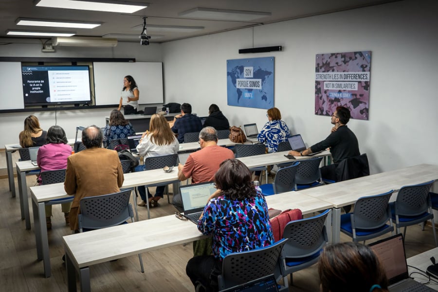 En la foto aparecen los y las docentes participando del taller. Sentados frente a su computador personal en una sala mirando a la persona encargada de hacer el taller, quien muestra una presentación en una pantalla grande