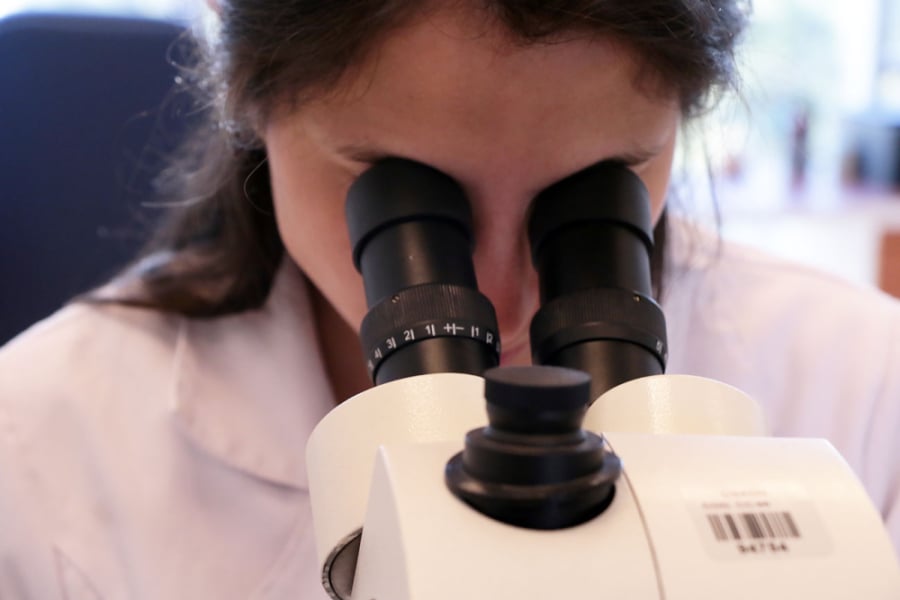 En la fotografía aparece una investigadora de delantal blanco, analizando un objeto desde un microscopio en el laboratorio.