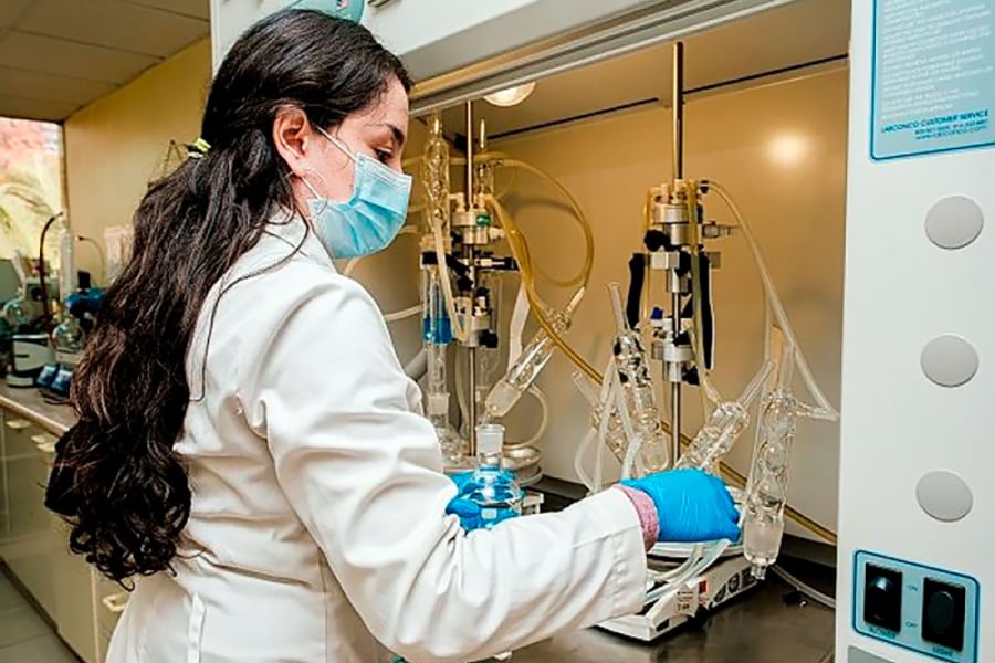 La imagen muestra una científica con bata blanca, guantes y mascarilla manipulando utensilios de laboratorio
