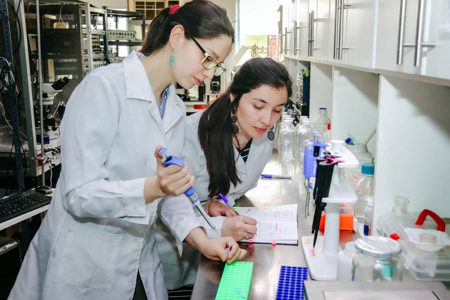 La imagen muestra a dos mujeres científicas con delantal blanco en un laboratorio: una manipulando instrumentos y la otra registrando lo observado