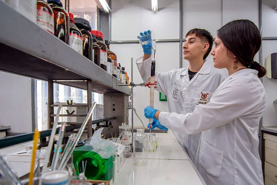 En la imagen aparecen dos estudiantes de la universidad trabajando en su laboratorio. Un hombre y una mujer que visten delantal blanco trabajan con pipetas y frascos.