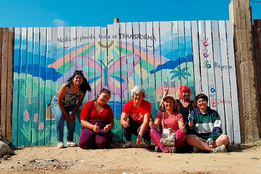 En la fotografía aparecen las estudiantes Usach y las pobladoras afectadas por los incendios de Quilpué.. El entorno es un mural de colores pasteles y un gran mariposa en el centro.