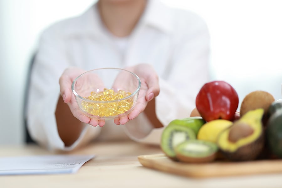 Person holding bowl of gel capsules beside fresh fruits, vegetables. 