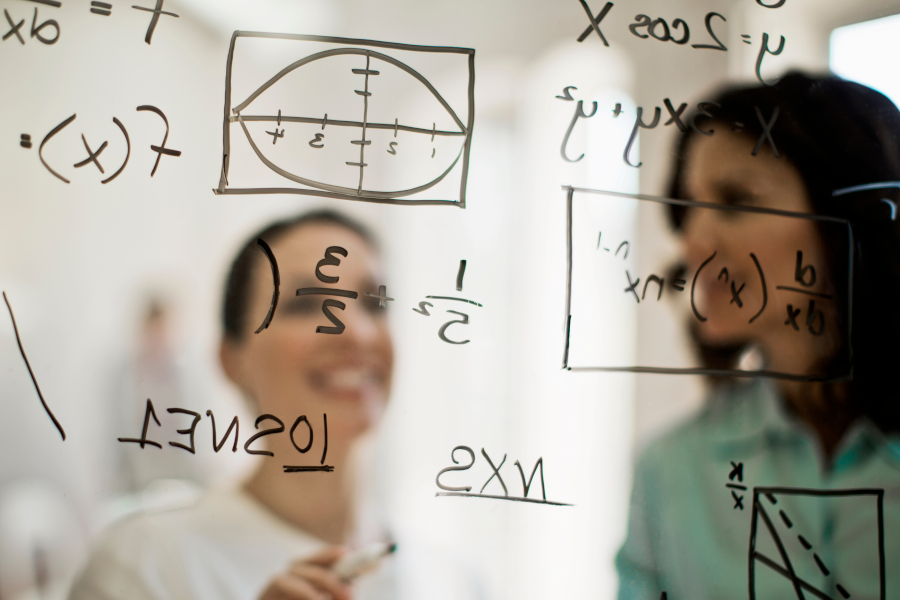 Ecuaciones matemáticas escritas en un cristal con dos mujeres al fondo.