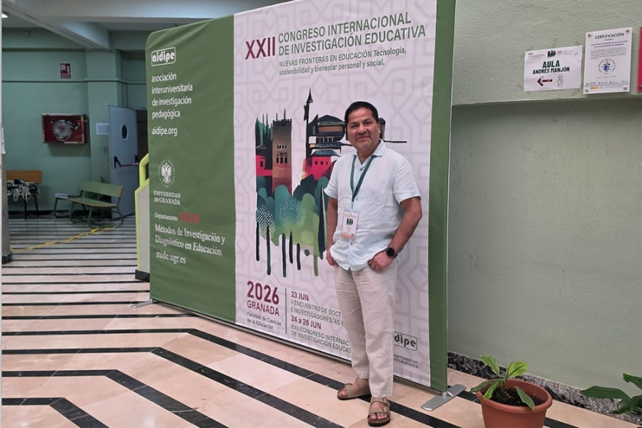 La foto muestra al investigador posando con las manos en los bolsillos, frente al pendón del evento