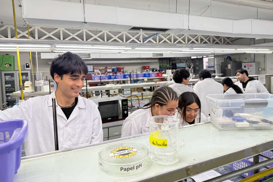 La imagen muestra a estudiantes en un laboratorio, usando bata blanca y manipulando elementos