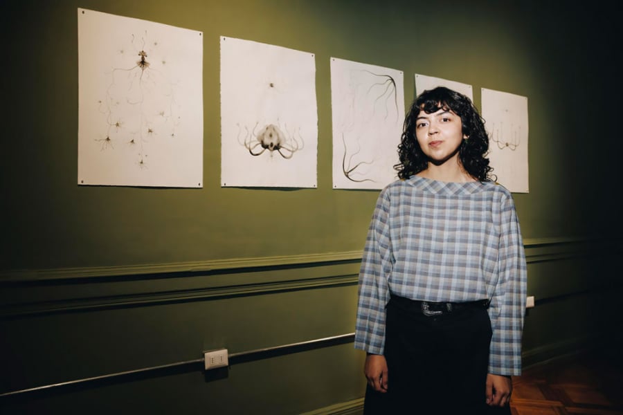 La foto muestra a la artista visual posando frente a una de sus exposiciones. De pie con una blusa a cuadros y pantalones negros, de fondo la pared verde y cinco de sus obras
