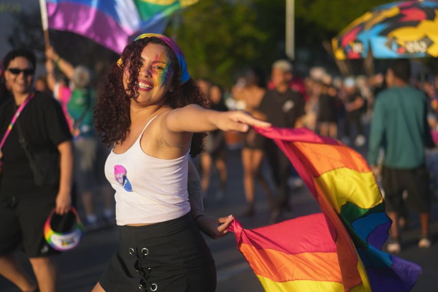 Una persona sonriente sosteniendo una bandera arcoíris del Orgullo en una manifestación al aire libre.