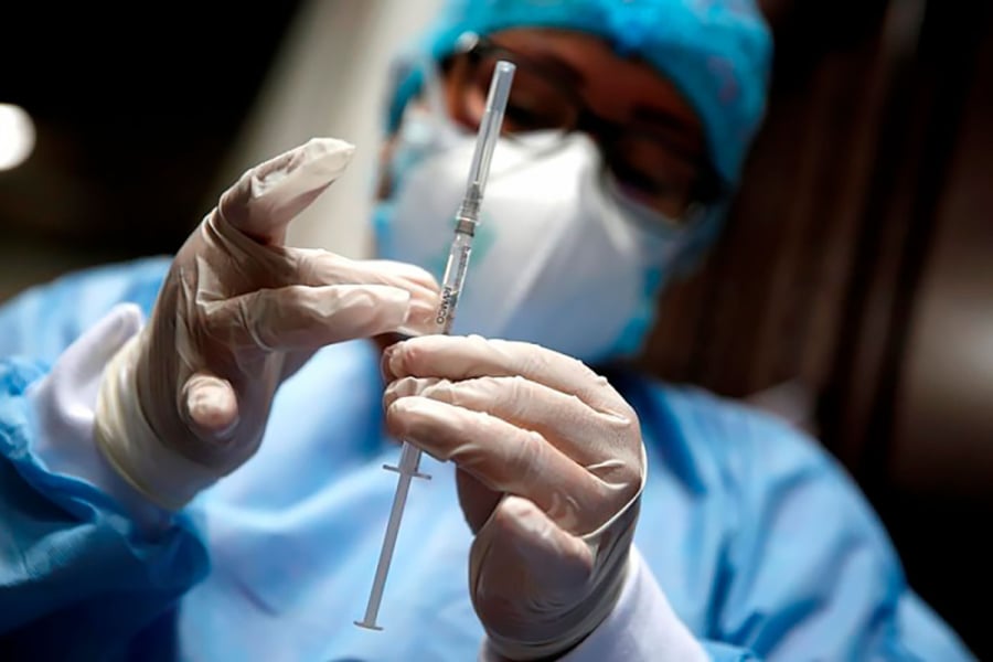 La imagen muestra a un médico sosteniendo una jeringa. Con guantes quirúrgicos, cofia, mascarilla y bata celeste 