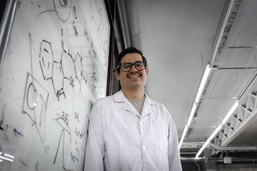 La imagen muestra al académico, de bata blanca, sonriente, apoyado en una pizarra de su laboratorio